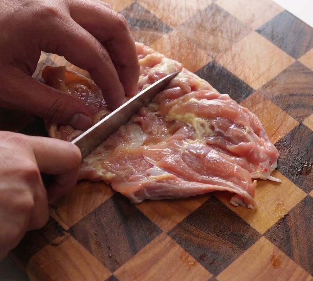 格子柄の木製まな板の上で、包丁の背を使って鶏もも肉を叩いて伸ばしている様子。