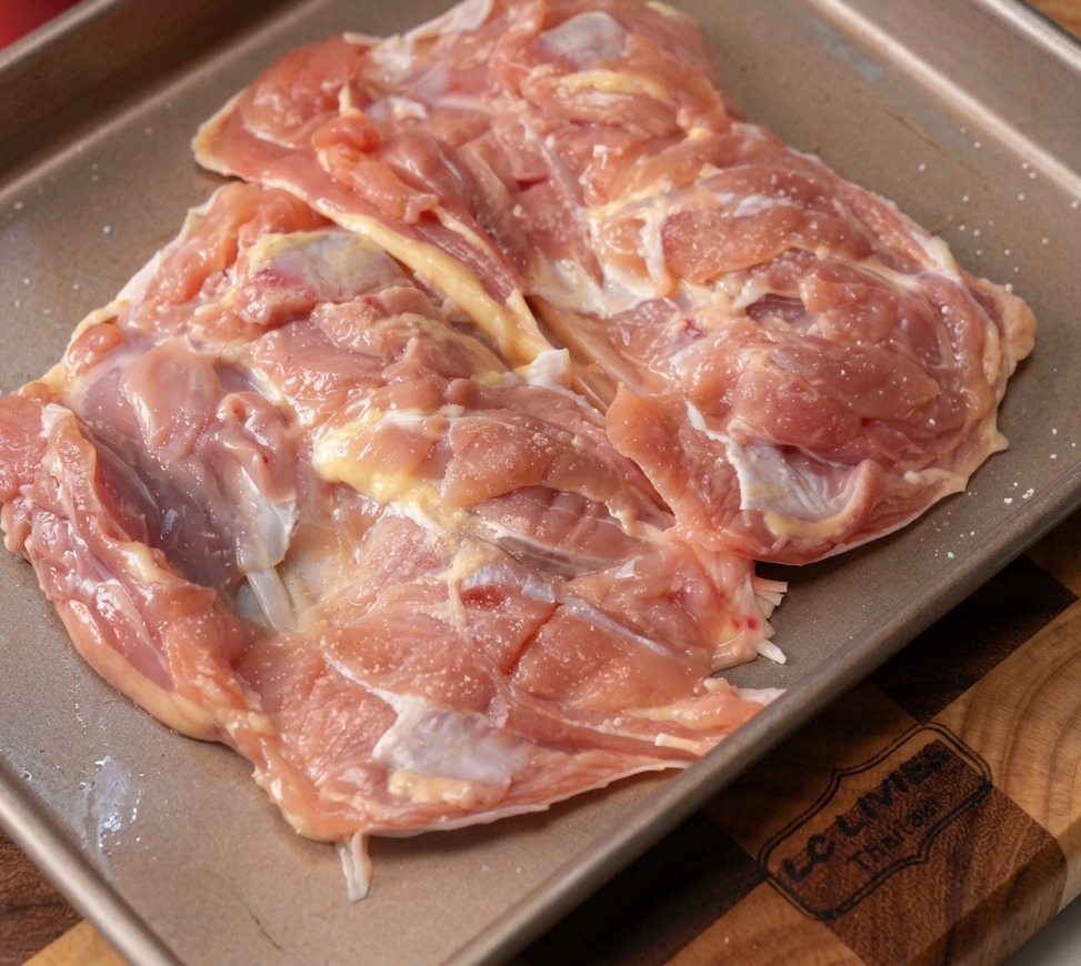バットに載った生の鶏もも肉に、みじん切りのにんにくと黒胡椒が振りかけられている様子。