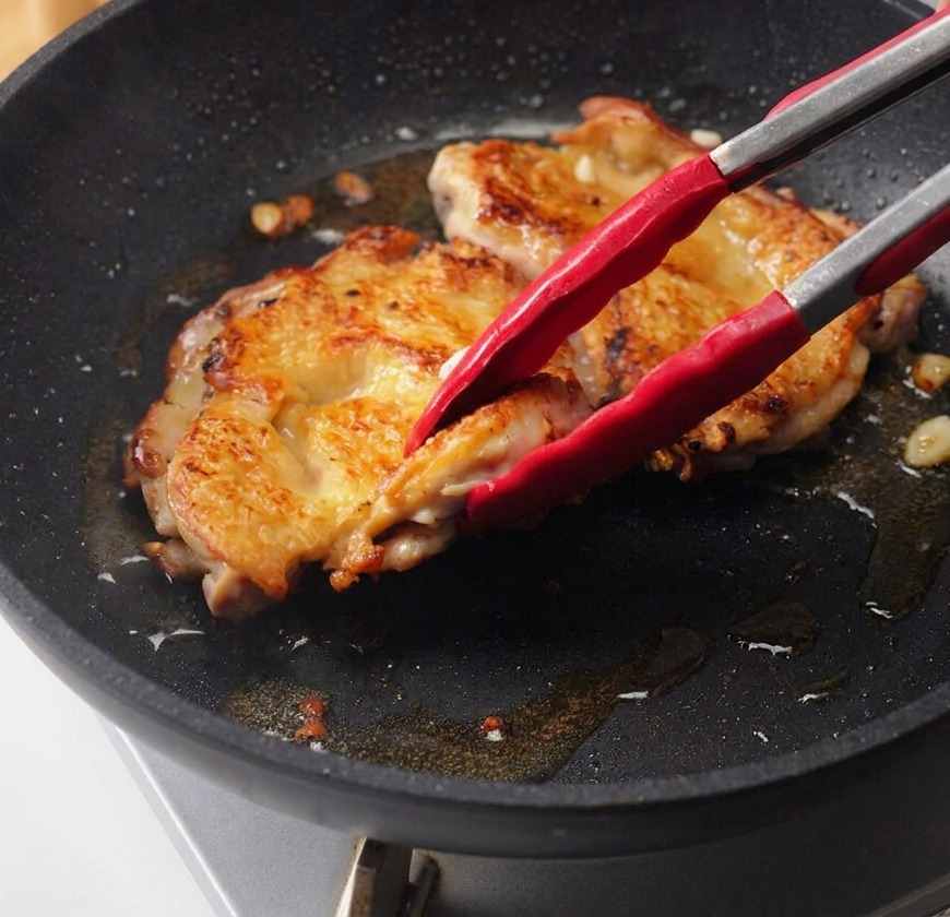 ガラスのボウルから、フライパンで焼いている鶏もも肉の上へ、濃い色の照り焼きタレを回しかけている手元。
