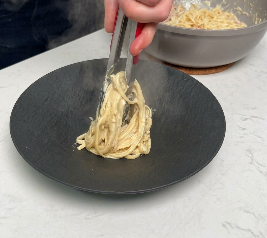 トングでパスタをくるくると巻きながら皿の中央に高く盛る様子