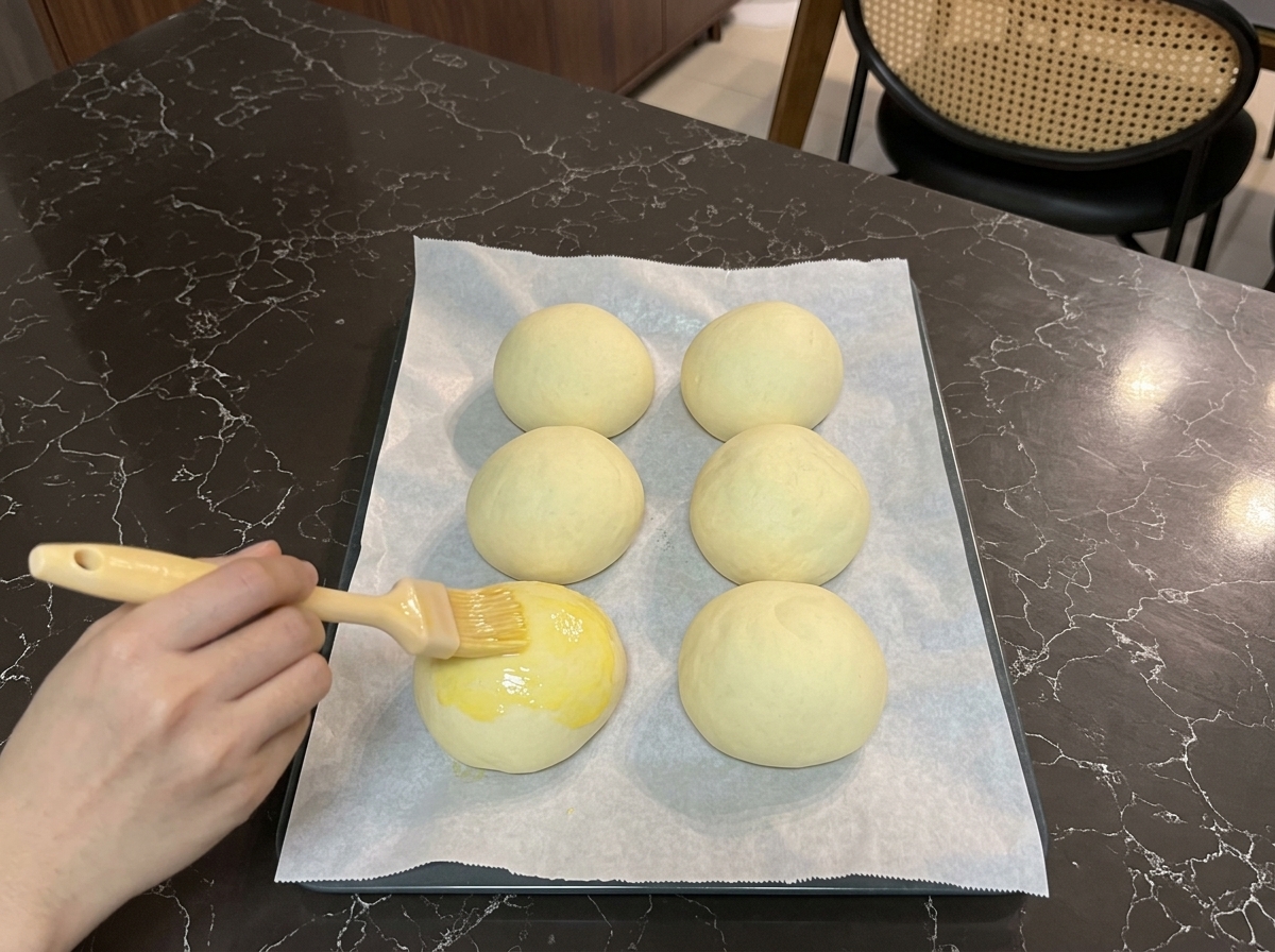 発酵が終わったバンズの表面に、ハケを使ってツヤのある黄色の卵液を塗っている手。