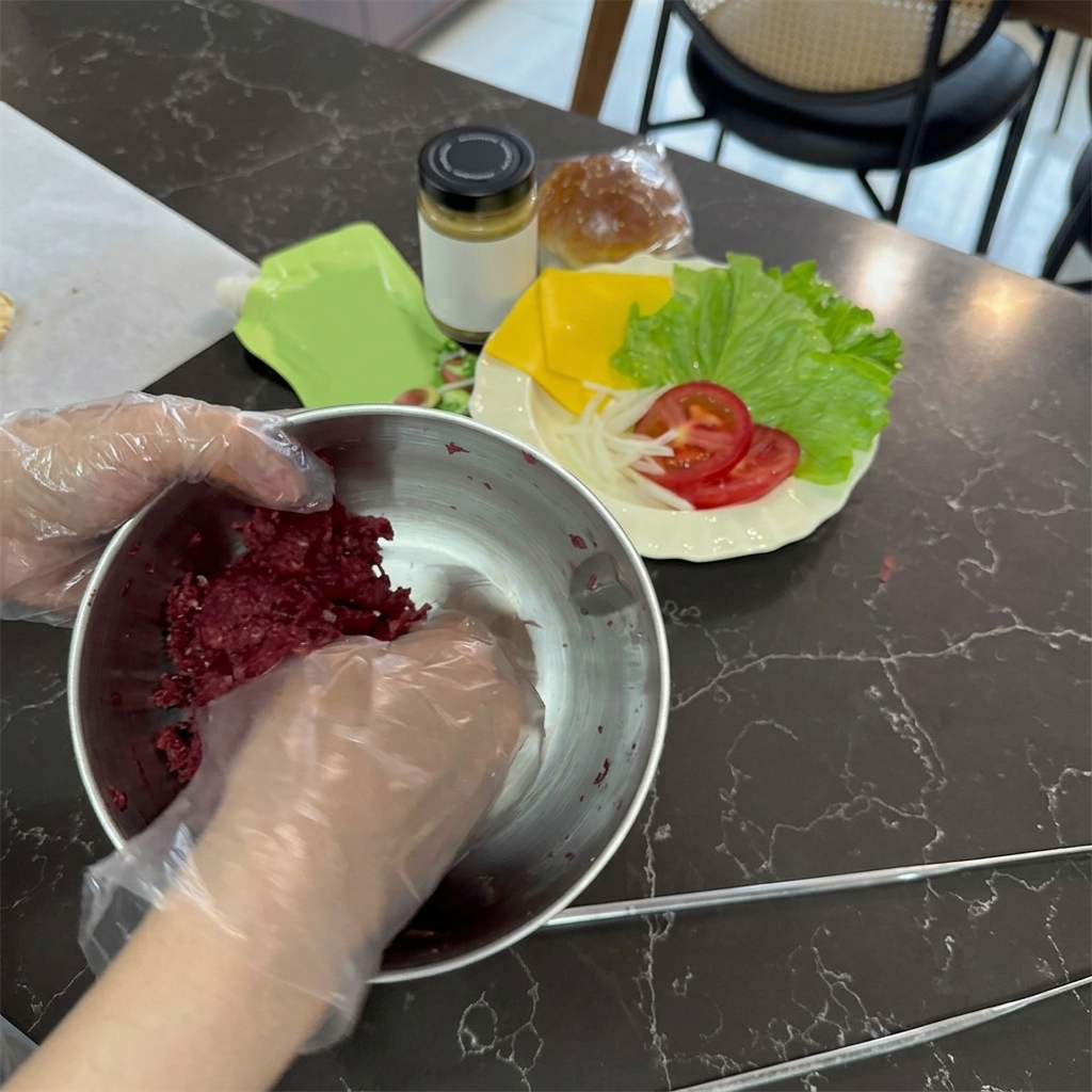 使い捨ての透明な手袋をした手で、ステンレスボウルに入った鮮やかな赤身のひき肉をこねている様子。