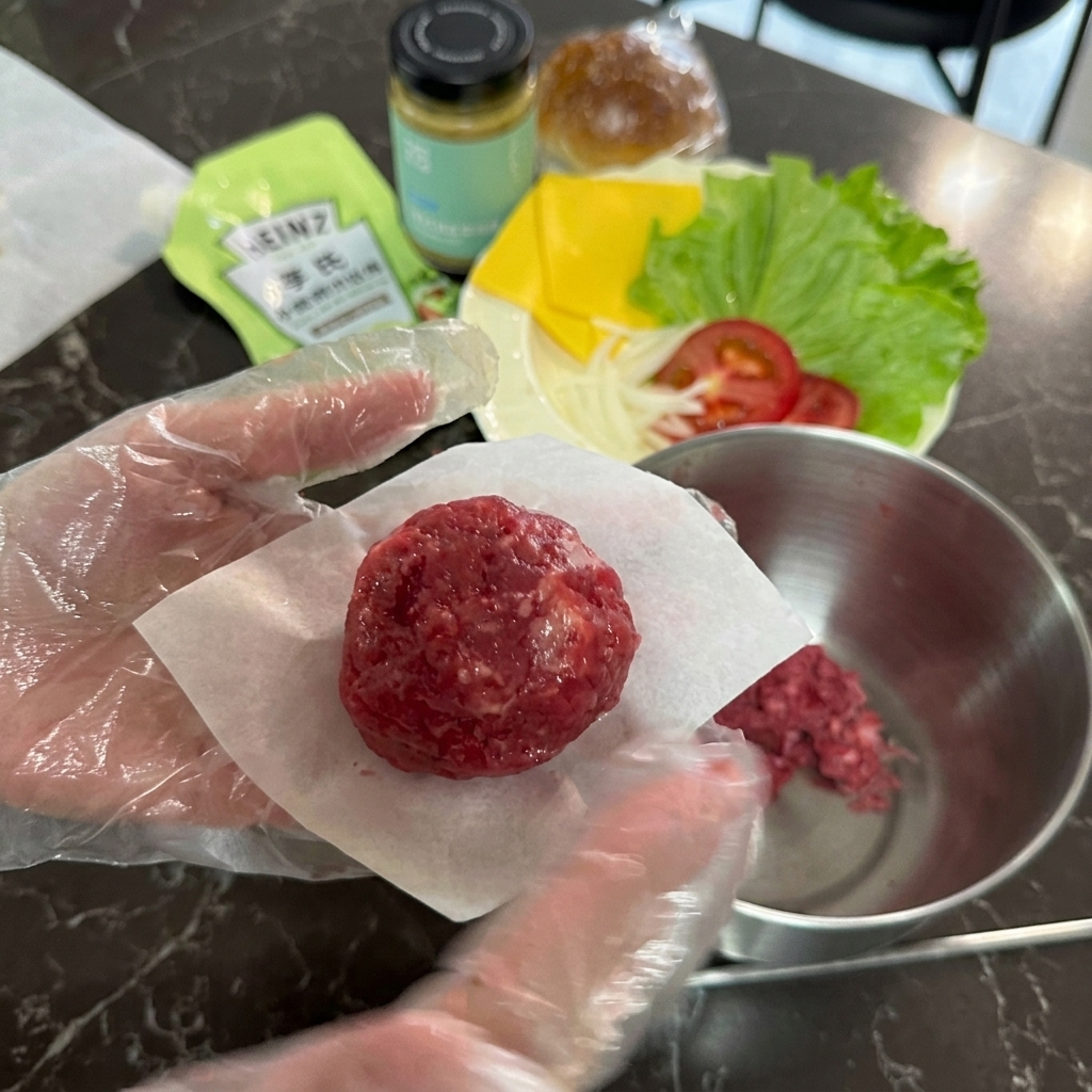 手袋をした手で、小さな四角いクッキングシートの上に生肉の丸いボールを置いている様子。