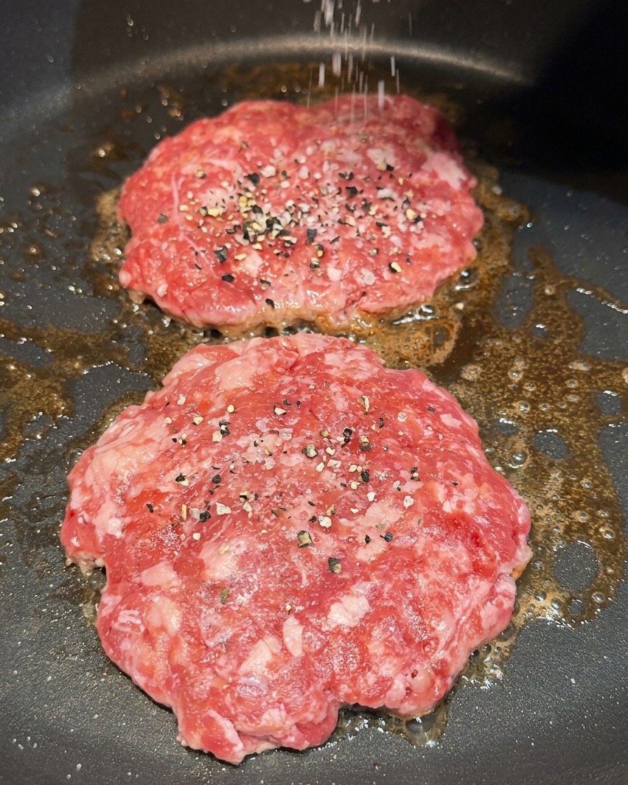 フライパンで焼いているビーフパティに、粗塩と黒こしょうを振りかけている様子。