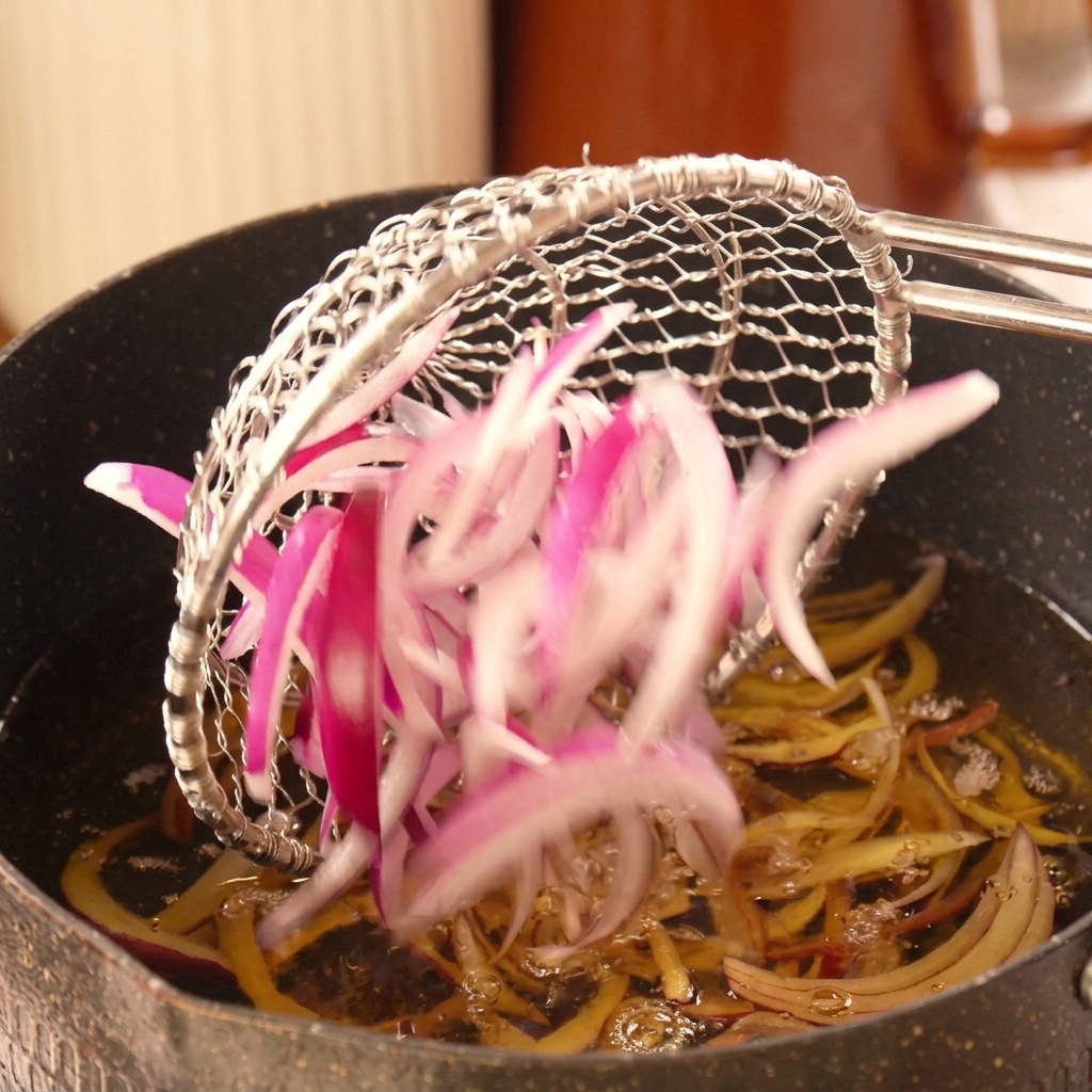 スライスした赤玉ねぎを、カス揚げを使って熱い油の中に静かに入れている様子。