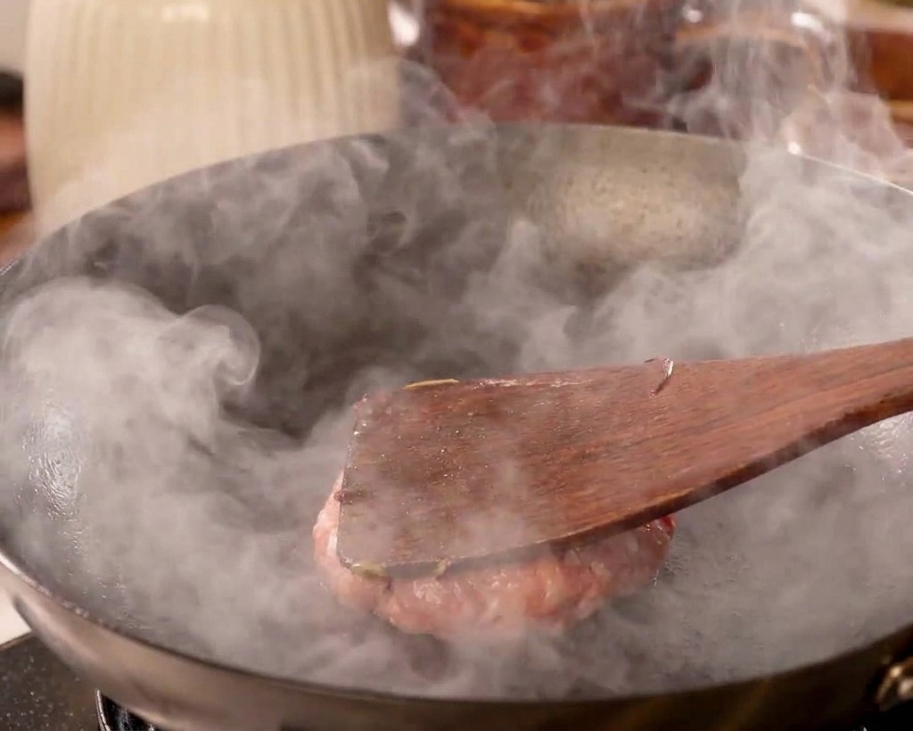 木製のヘラを使って、熱いフライパンの上で肉を力強く押しつぶしている様子。