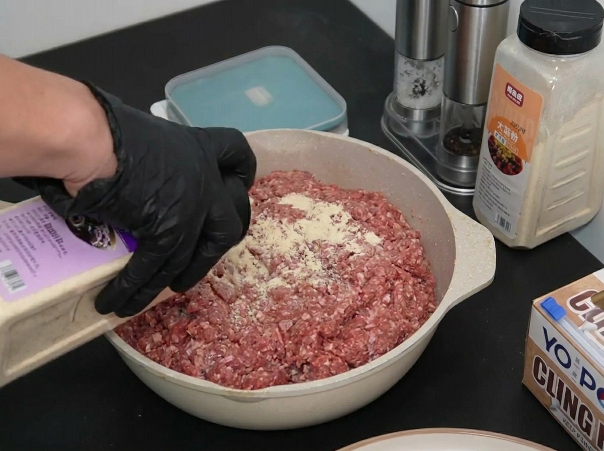 バットに広げた牛挽肉に、手袋をした手でオニオンパウダーを均一に振りかけている様子。
