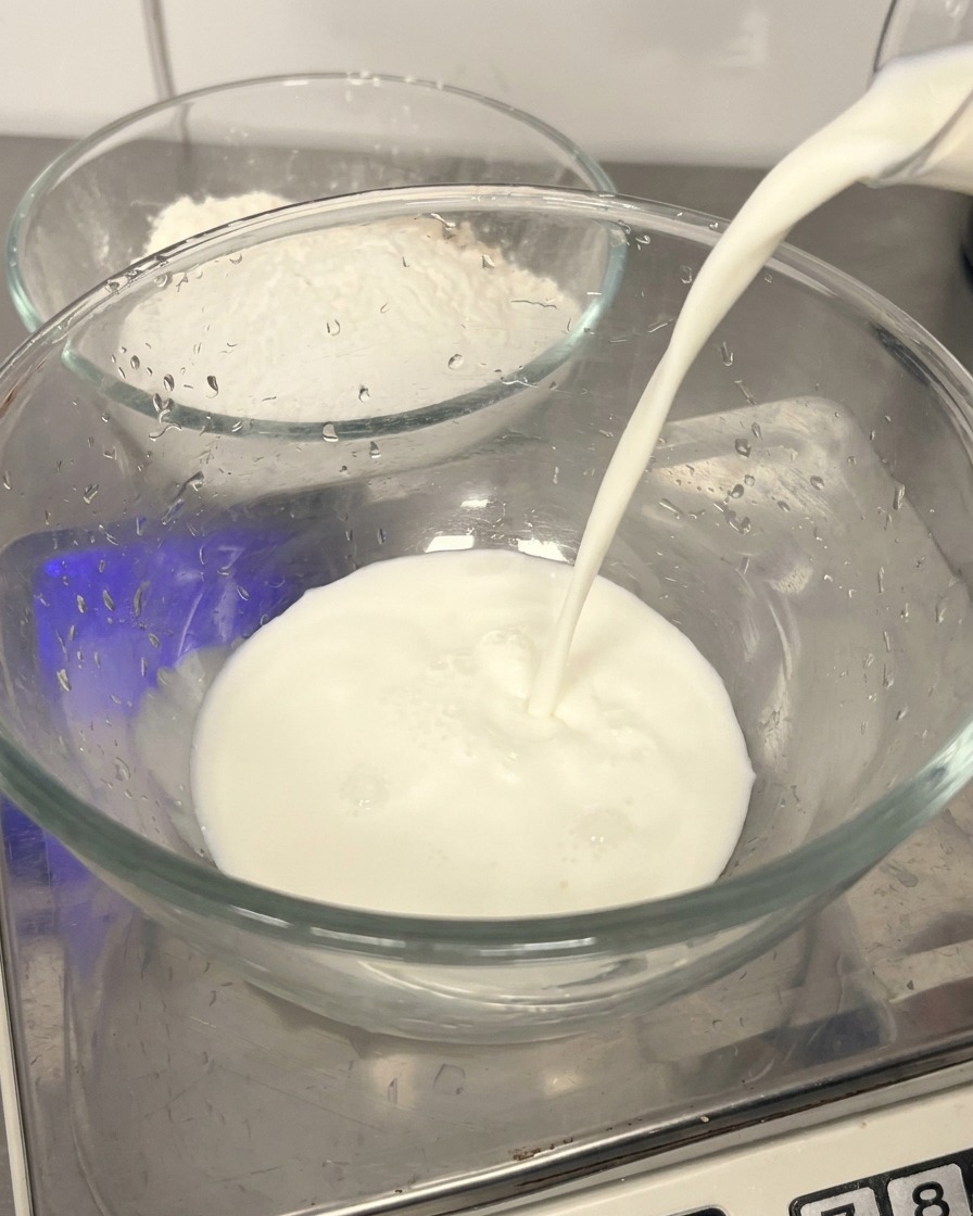 ボウルの背景に小麦粉が見える中、透明なガラスボウルに新鮮な牛乳を注いでいる様子
