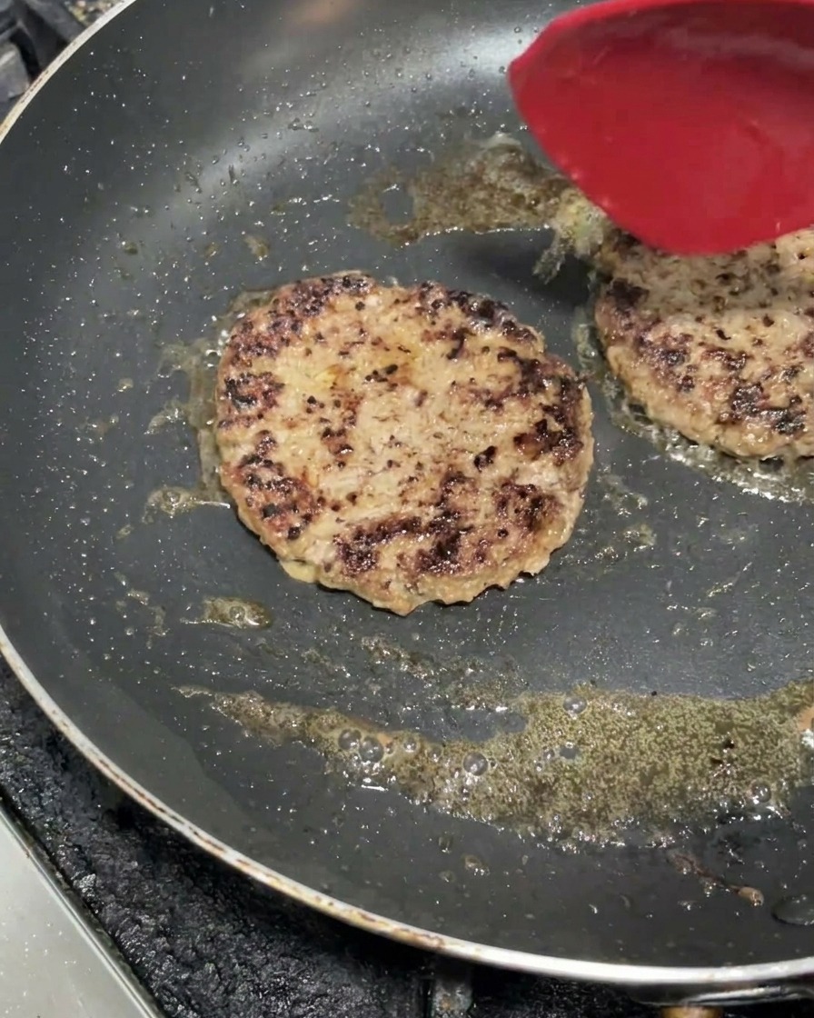 熱いフライパンでシズル感を出しながら焼ける2枚の肉厚パティと、その上に置かれた赤いヘラ