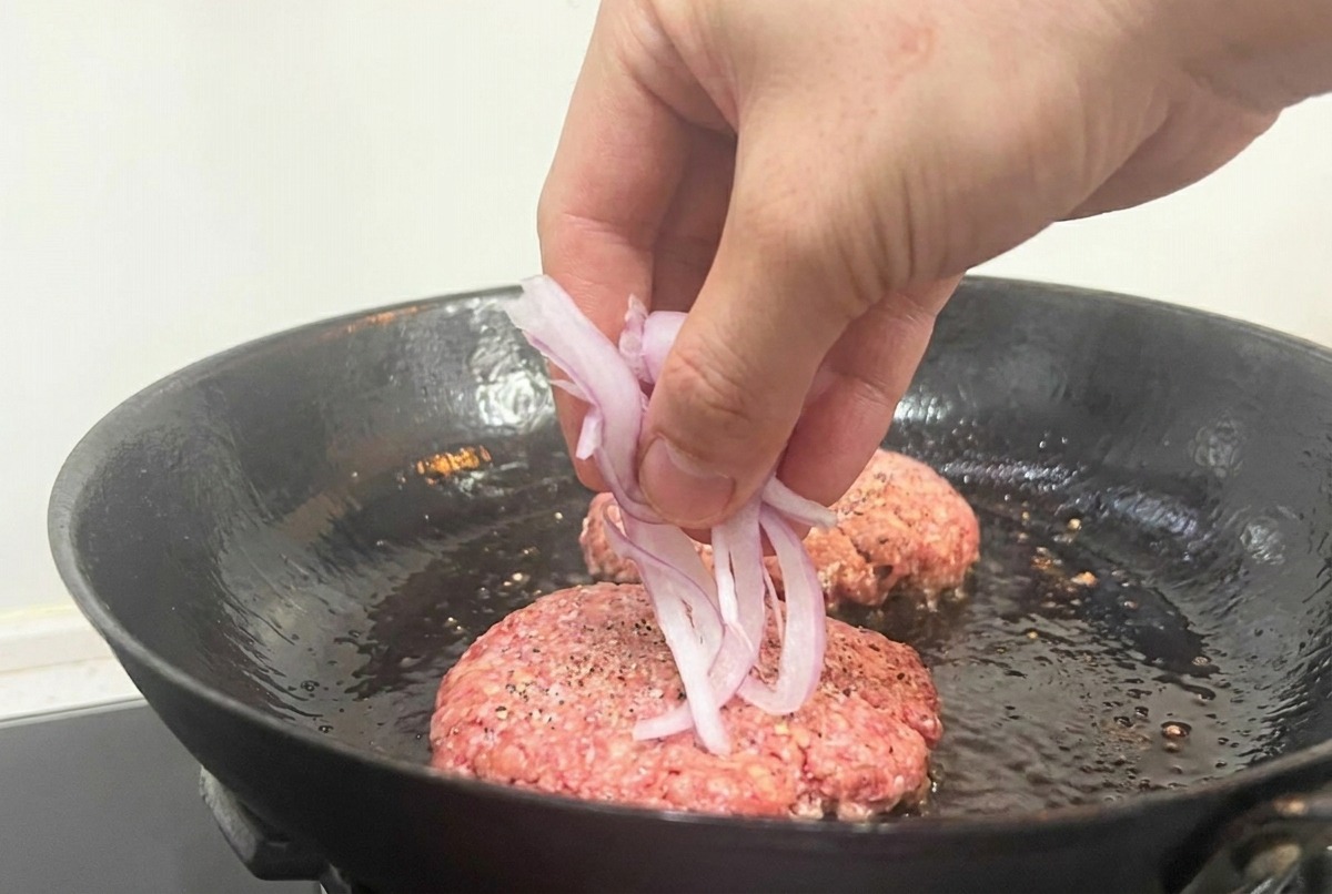 焼いている最中のパティの上に赤玉ねぎのスライスをのせる様子