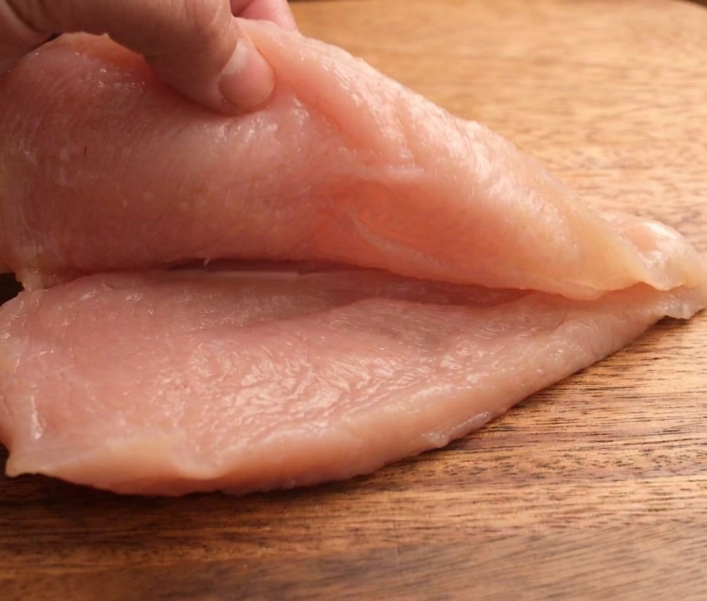 木製のまな板の上で、生の鶏むね肉を水平に包丁で切り、薄い2枚に分けている様子。