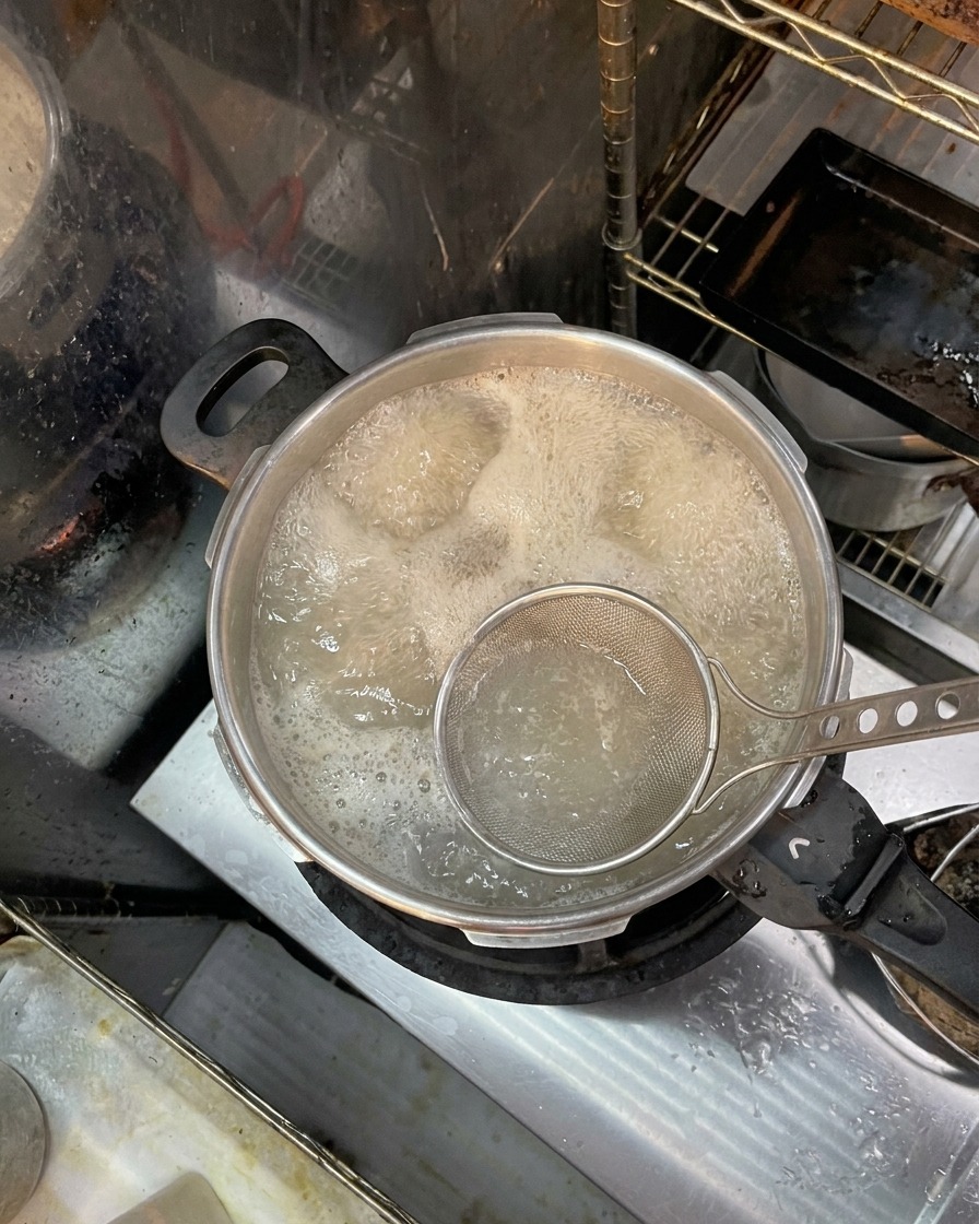 沸騰した鍋の表面に浮いた茶色い泡やアクを、網じゃくしですくい取っている様子。