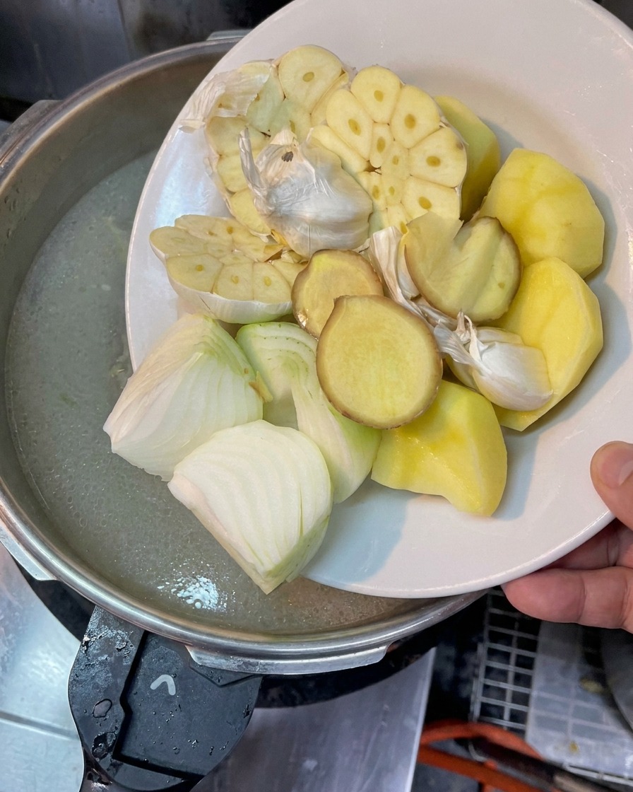 半分に切ったにんにく、玉ねぎ、生姜、じゃがいもを煮立ったスープに加えようとしている様子。