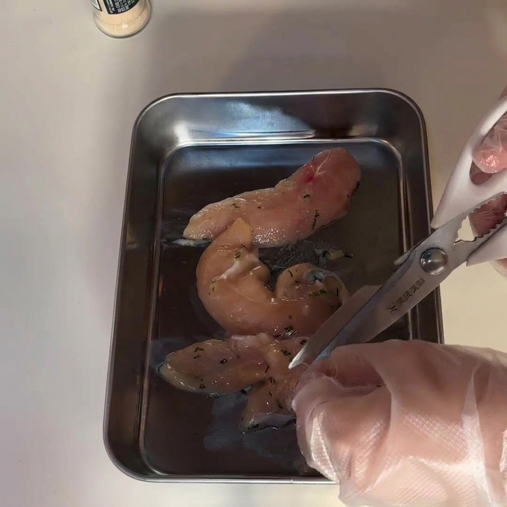 ステンレスのトレイの上で、キッチンのハサミを使って生の鶏むね肉をカットしている様子。
