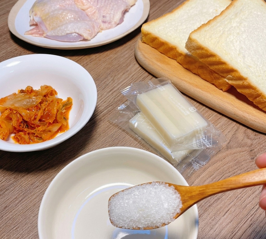 材料のキムチやパン、鶏肉が並ぶ中、白いボウルの上で砂糖をすくう木のスプーン。