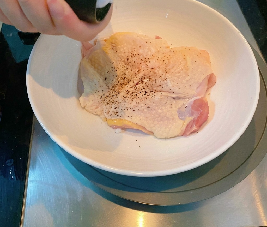 白い陶器のボウルに入った生の鶏もも肉に、ペッパーミルで胡椒を振っている様子。