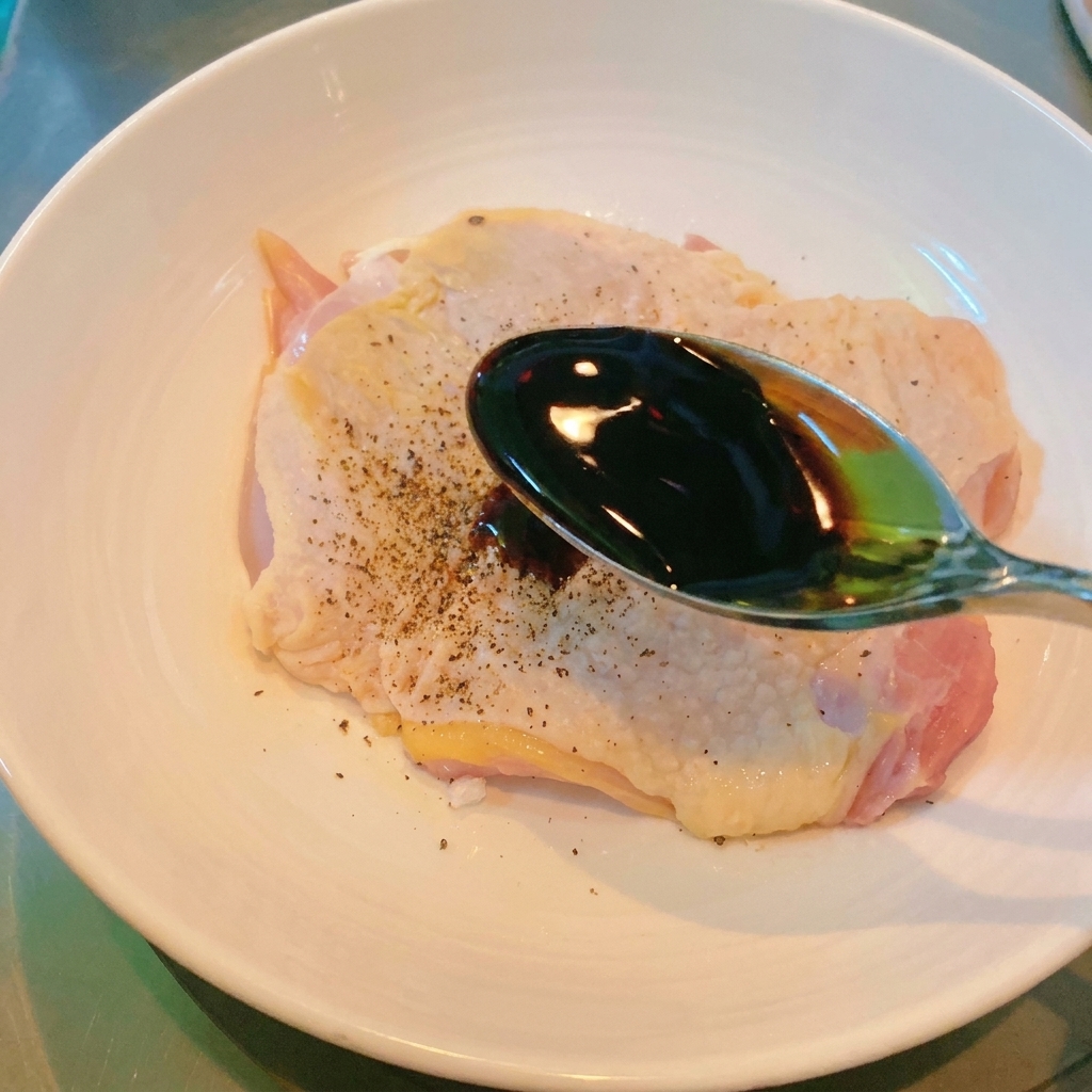 ボウルに入った下味済みの鶏もも肉に、スプーンで濃い色の醤油をかけている様子。