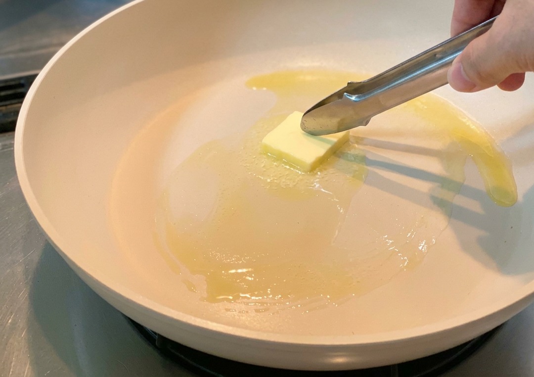 加熱されたフライパンの中で、四角いバターが溶け出し、トングで動かされている様子。