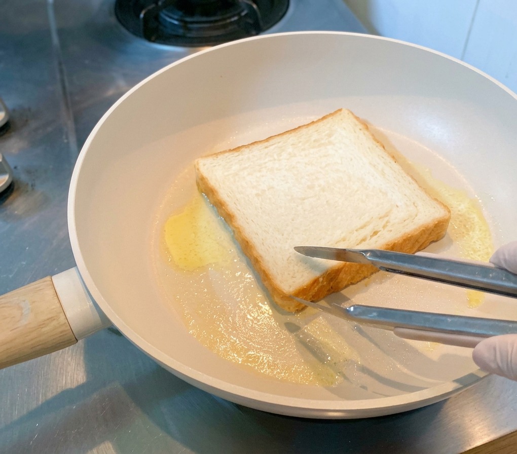 バターが溶けたフライパンで、トングに押さえられながら焼かれている食パン。