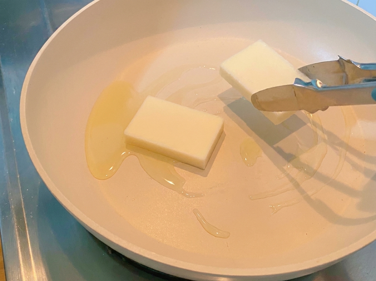 加熱されたフライパンに、厚みのある長方形の白いトックがトングで並べられている様子。