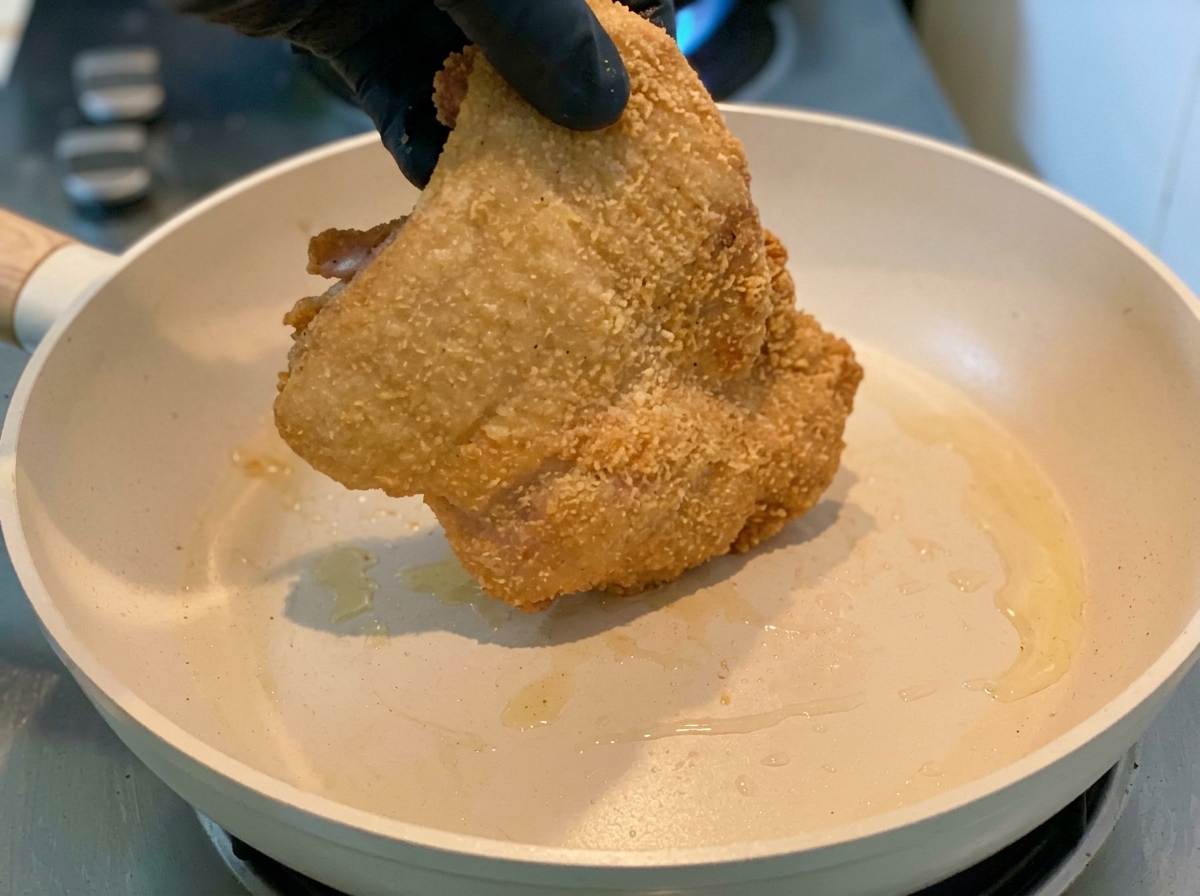 手袋をした手で、片栗粉を纏った鶏もも肉を熱いフライパンに入れている様子。