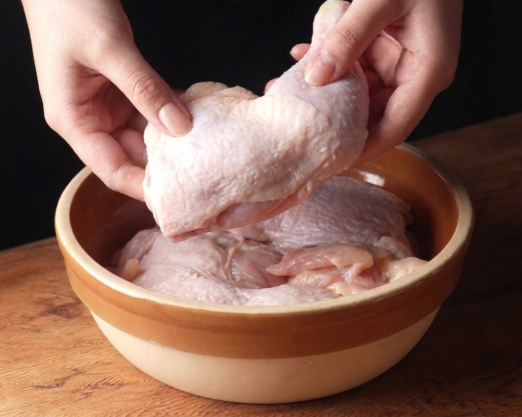 木製の台の上にあるベージュのセラミックボウルに、生の鶏もも肉を入れる様子。