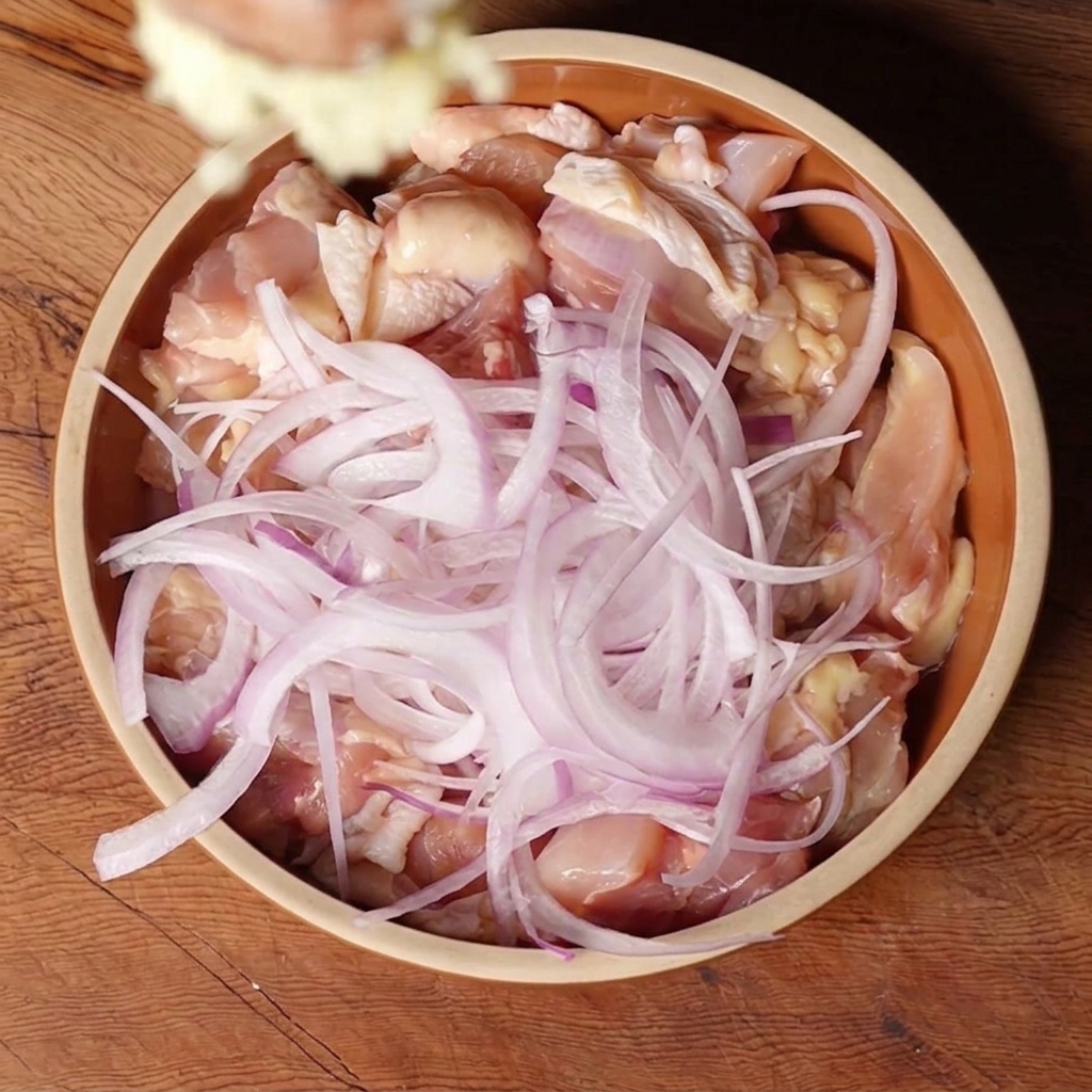 カットした鶏肉と赤玉ねぎが入ったボウルに、おろしにんにくを投入しているシーン。