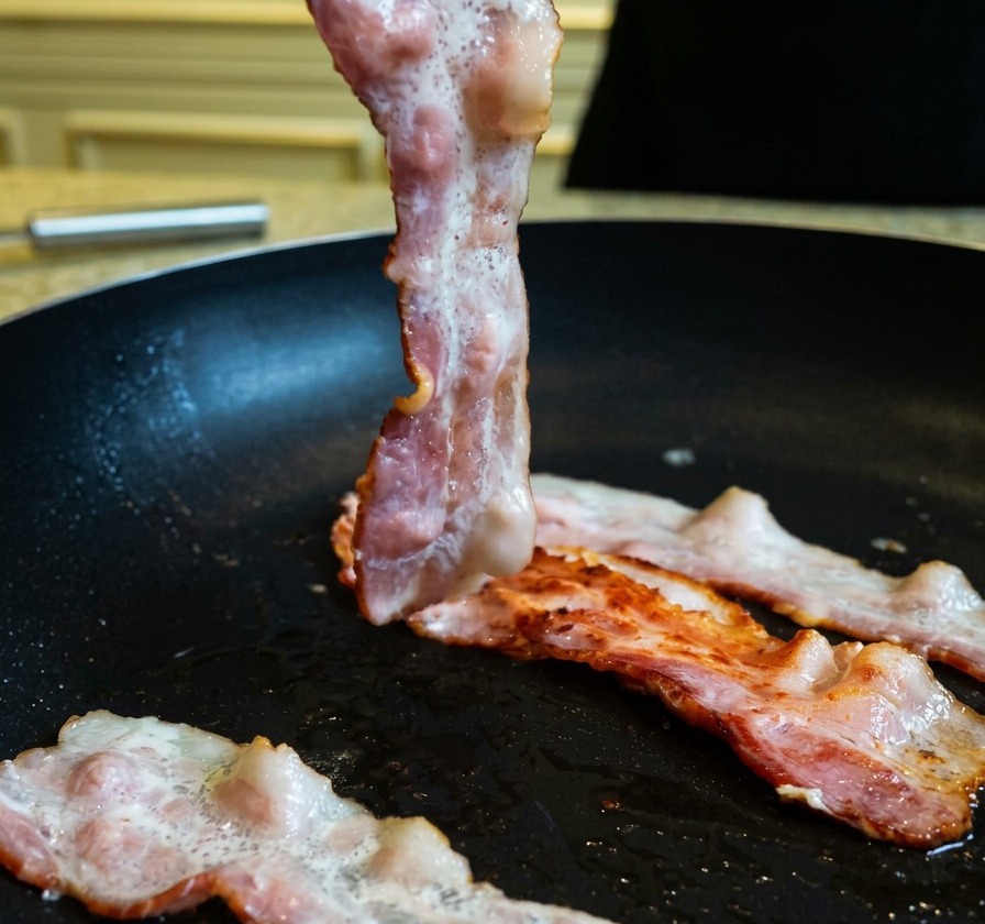 トングを使い、焼き色がついたベーコンをフライパンで裏返している様子。