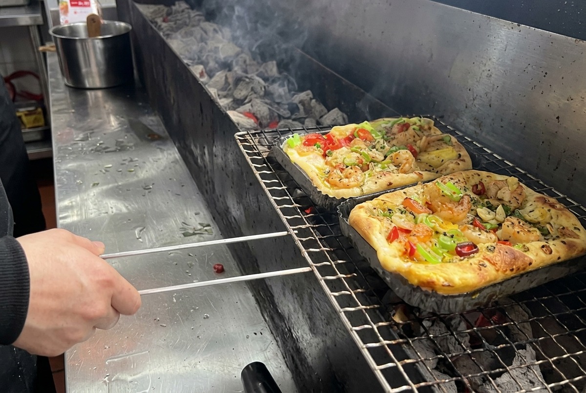 プロの厨房で、エビやミックス野菜をトッピングした長方形のピザを細長い炭火グリル網にのせている様子。