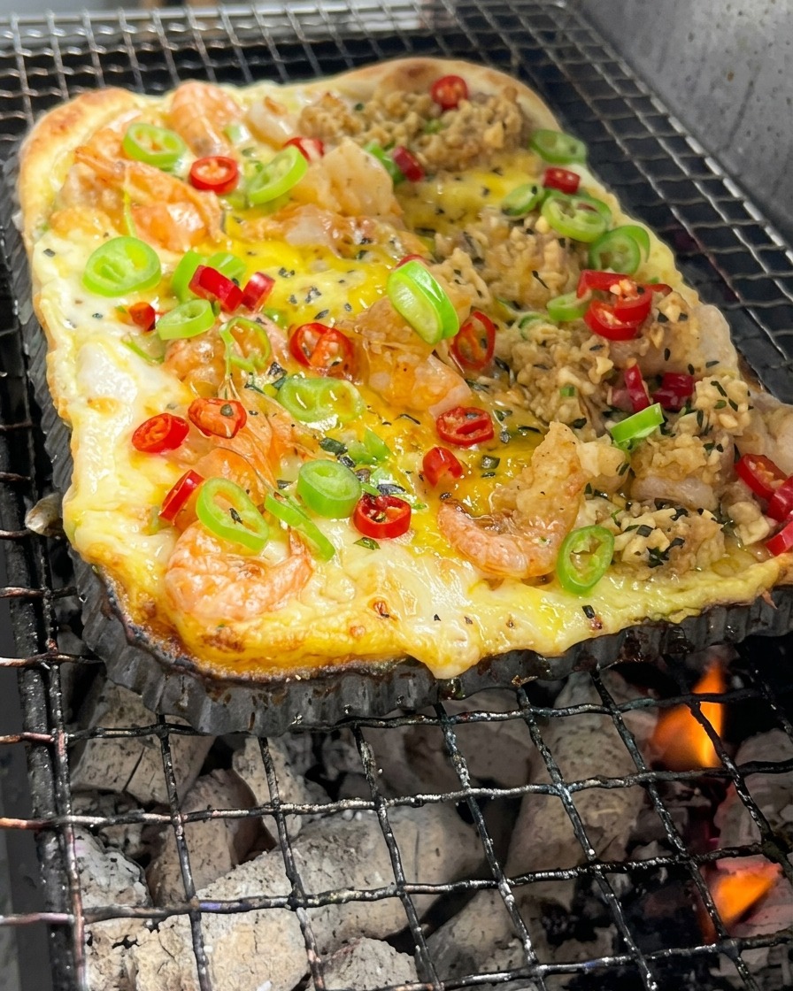 赤く輝く炭火の上で、エビや野菜がのった長方形のピザ生地が焼かれている様子。