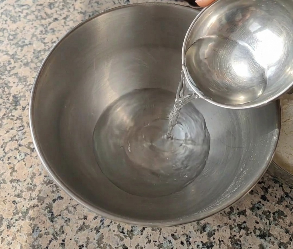 グラニットの作業台の上で、小さな金属製のボウルから大きなステンレス製のミキシングボウルへ水が注がれている様子。