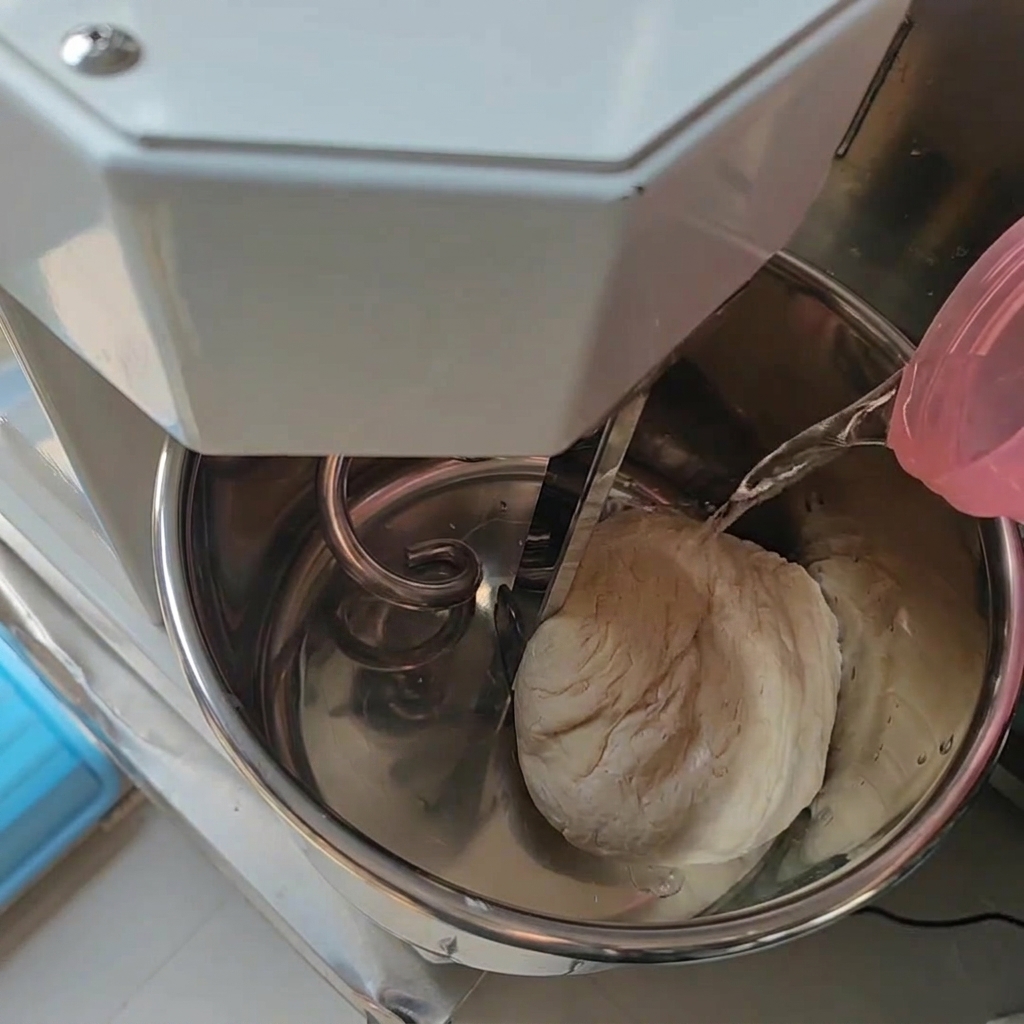 すでに生地の塊が入っているステンレス製のミキサーボウルに、ピンクの容器から水が注がれている様子。