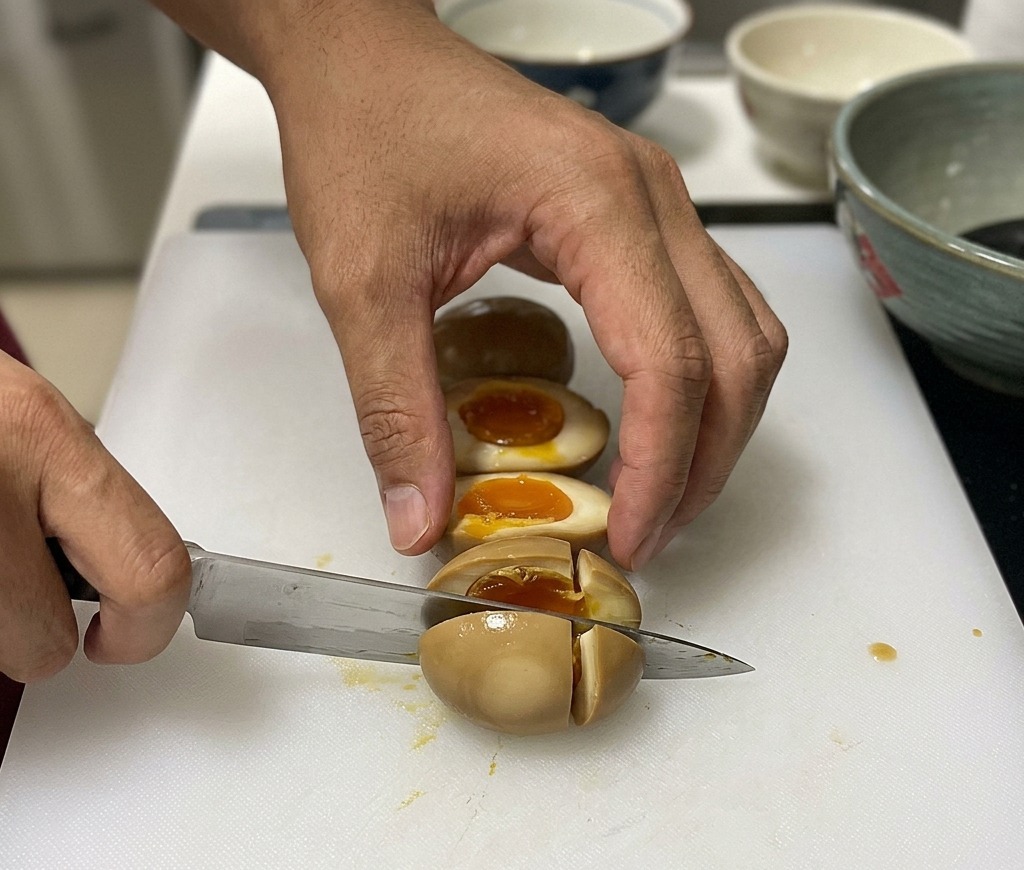 白いまな板の上で、茶色く色づいた半熟味玉を包丁で半分に切っている様子。中からとろりとした黄身が見える。