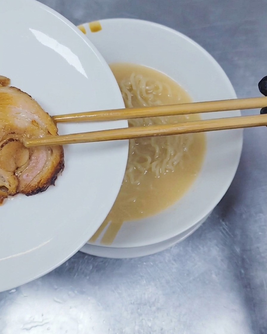 箸を使って、ラーメンの麺の上に丸く成形されたチャーシューを一切れそっとのせている様子