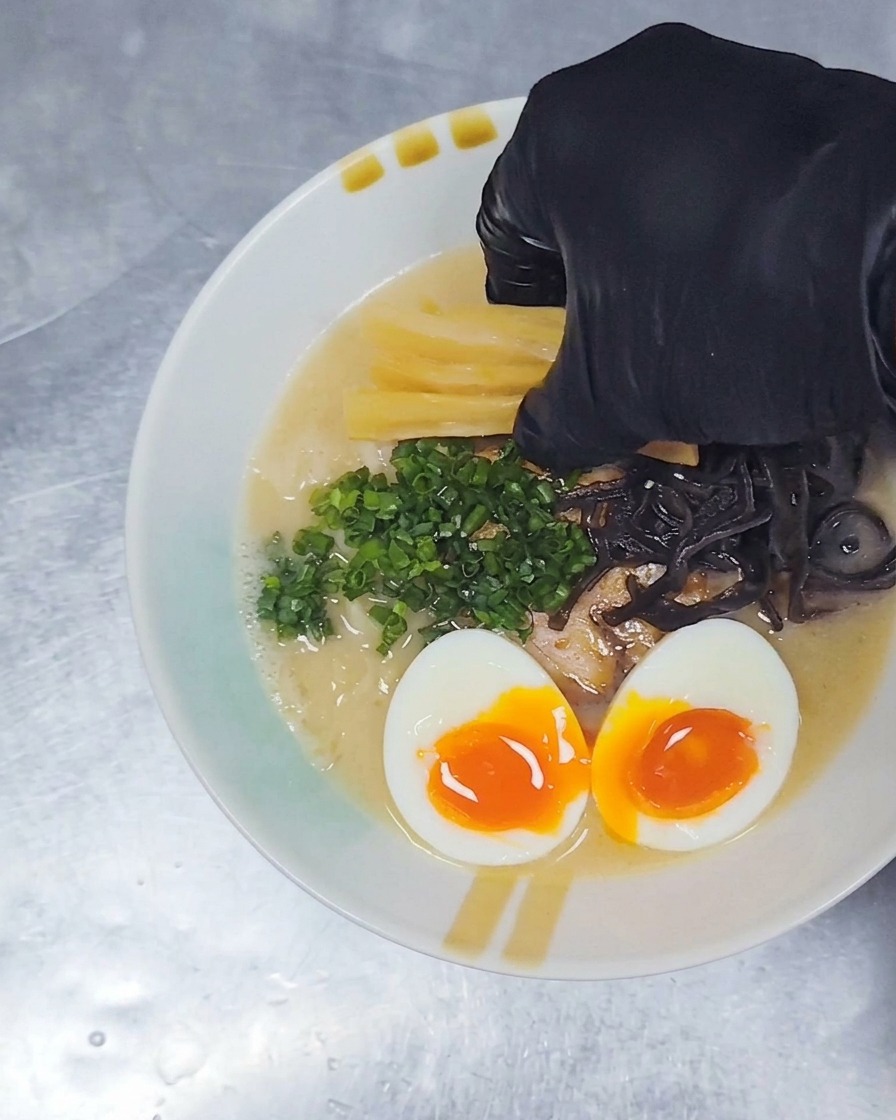 チャーシュー、卵、ネギなどがのったラーメンに、仕上げとして黄色いメンマを箸で添えている様子