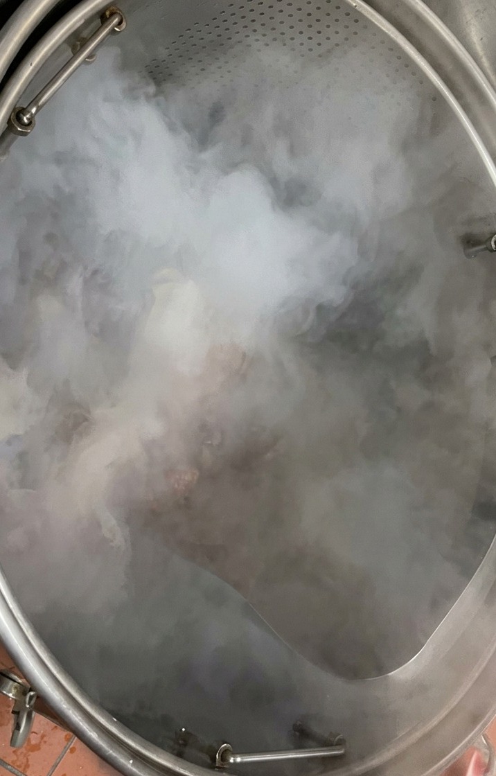 湯気が立ち上るステンレス製の深い寸胴鍋に、生の豚骨をトレイから投入している様子。