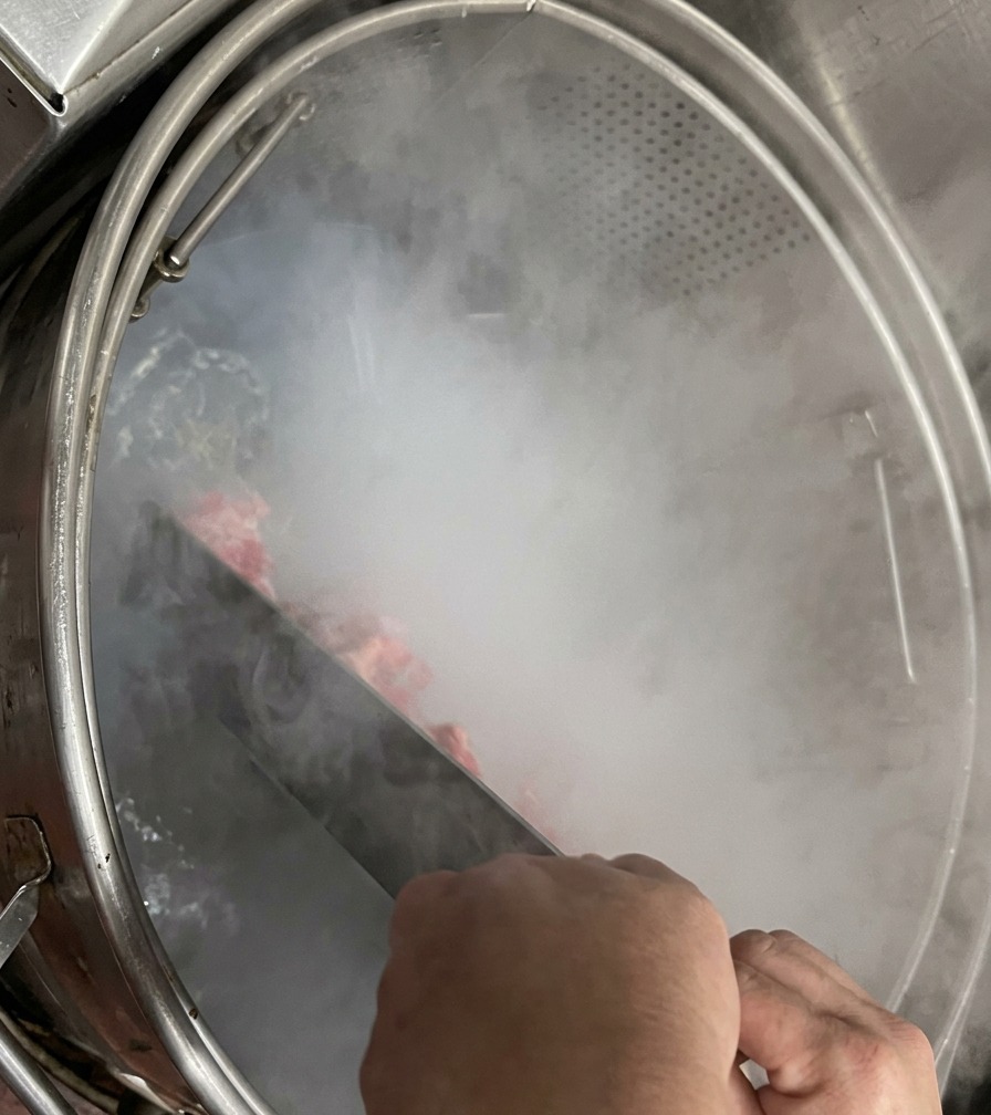 湯気が立ち上る大きな鍋の中で、大きな金属製のヘラを使って豚骨をかき混ぜている手元。