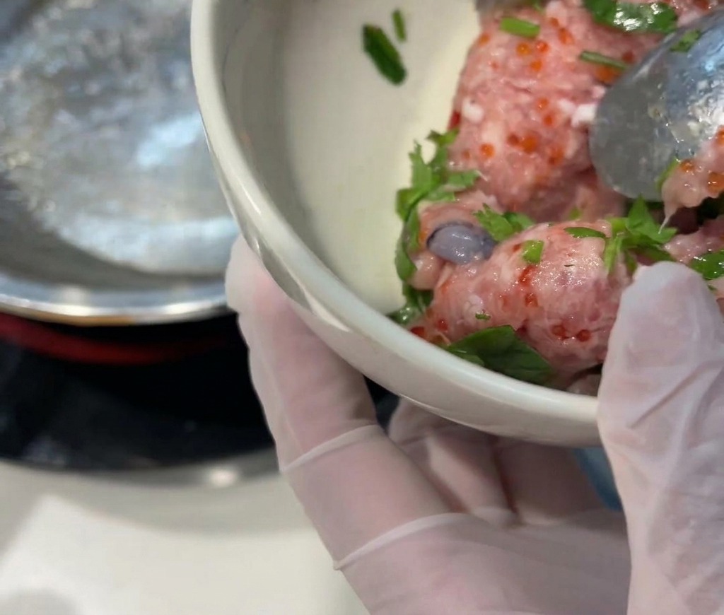 透明な手袋をはめた手が、生のピンク色のひき肉、鮮やかなオレンジ色のとびっこ、緑色のパクチー、そして新鮮なエビが入った小さな白いボウルを持っている様子。