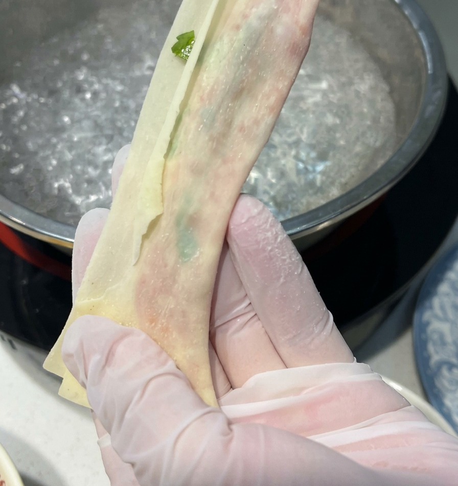 手袋をはめた両手が、沸騰したお湯が入った鍋の上で、餡を包んだワンタンの皮を細長く半透明なリボン状に伸ばしている様子。
