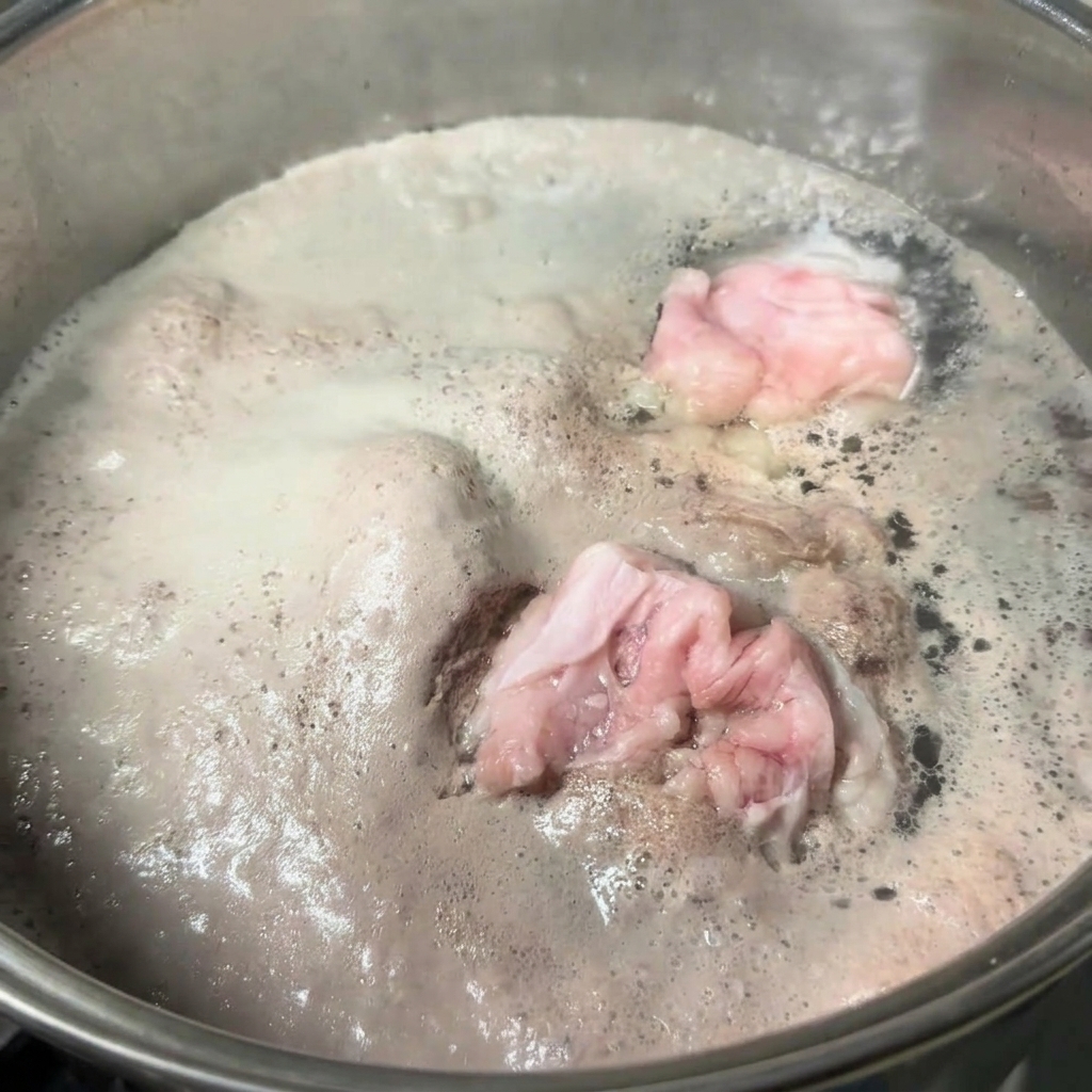 沸騰したお湯の中で豚骨が煮込まれ、表面に灰色の厚いアクの層が浮かんでいるステンレス鍋。