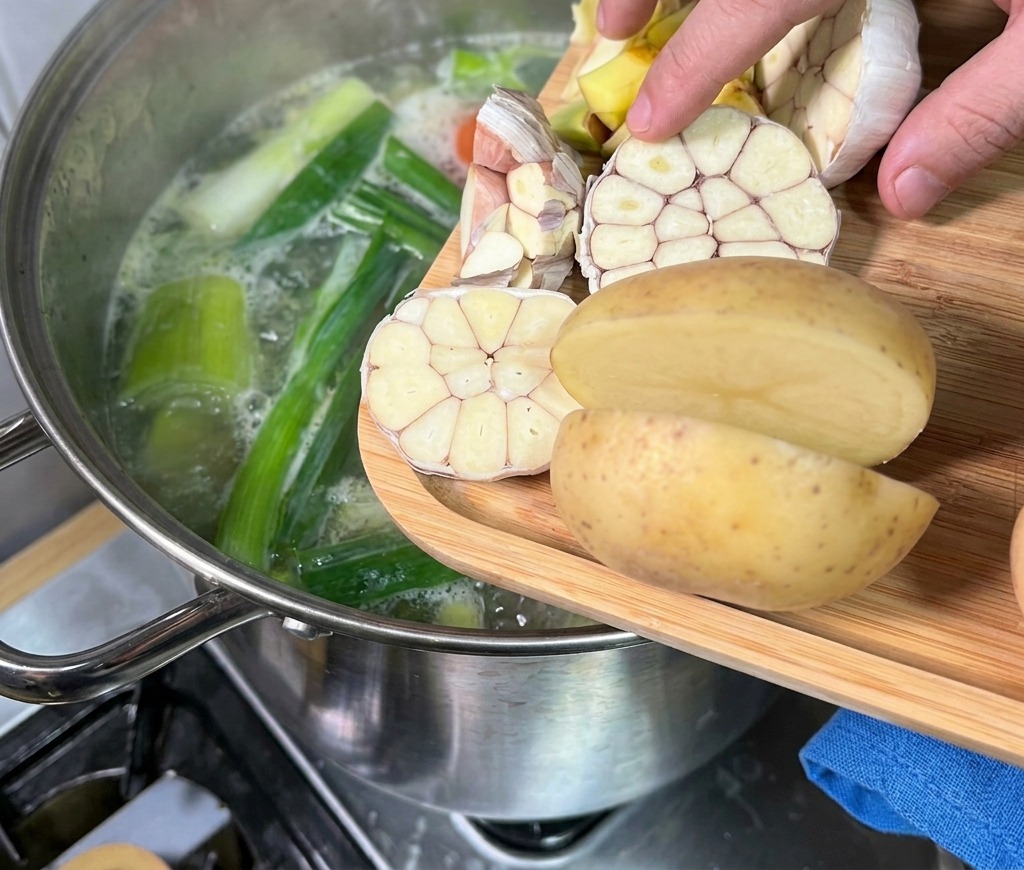 木製のまな板から、半分に切ったにんにくとじゃがいもを、湯気の立つ豚骨スープの鍋に手で投入している様子。