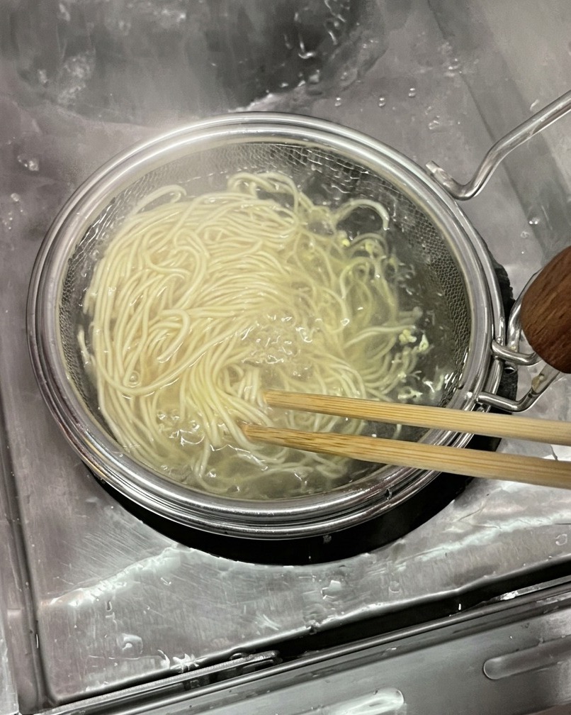 円形のメッシュ製テボ（振りザル）を使い、沸騰したお湯から細い生の小麦麺の束を木箸でほぐしながら引き上げている様子。