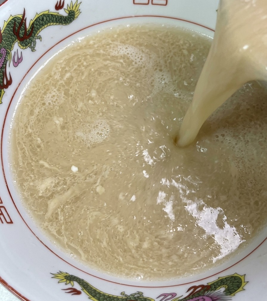 調味タレとラードが入った温かいラーメン丼に、お玉から湯気立つ乳白色の豚骨スープが注がれ、シルキーな質感が広がっている様子。