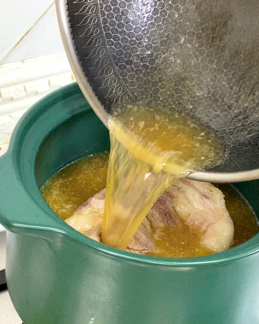 焼きつけた豚骨が入った緑色の土鍋に、フライパンから旨味の詰まったスープと油を注ぎ入れる様子。