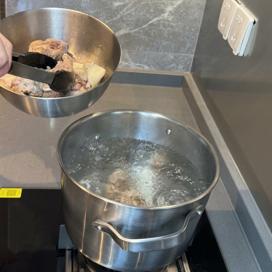 下茹で済みの豚骨が入ったボウルを、沸騰したお湯の入った鍋の上に持っている様子。