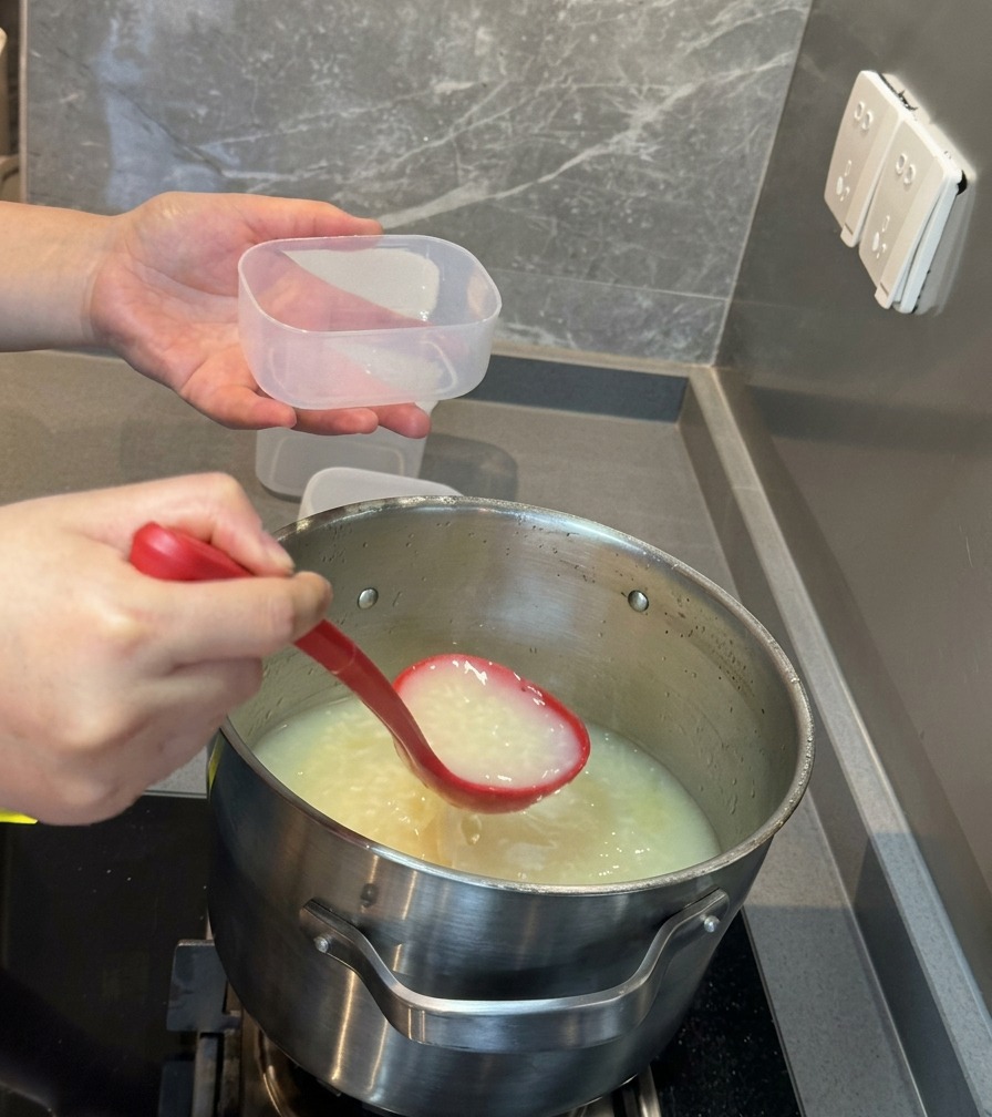 濃厚で真っ白な豚骨スープを、赤いお玉で透明なプラスチックの保存容器に注ぎ入れている様子。