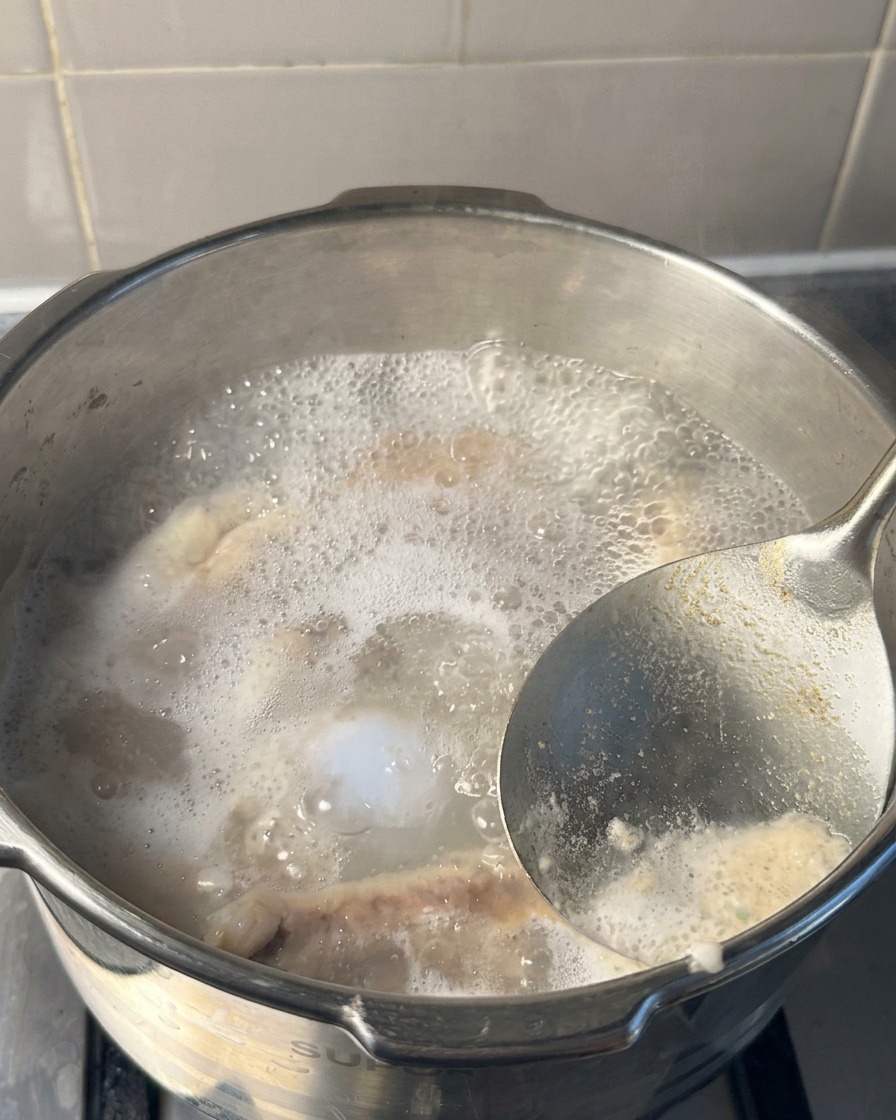 圧力鍋の中で沸騰するお湯の表面から、金属製のおたまを使って白やグレーの泡をすくい取っている様子
