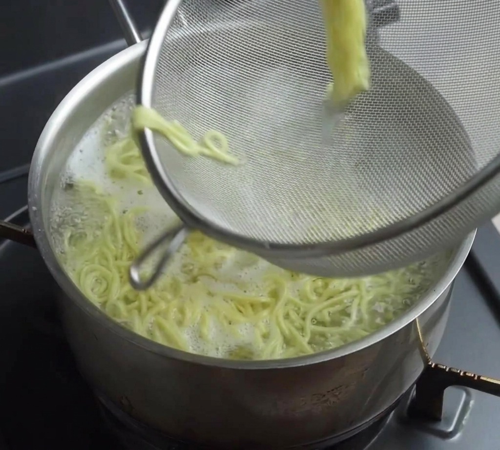 湯気が立ち上る鍋から、メッシュの湯切りザルで茹で上がった黄色い中華麺を引き上げている様子。