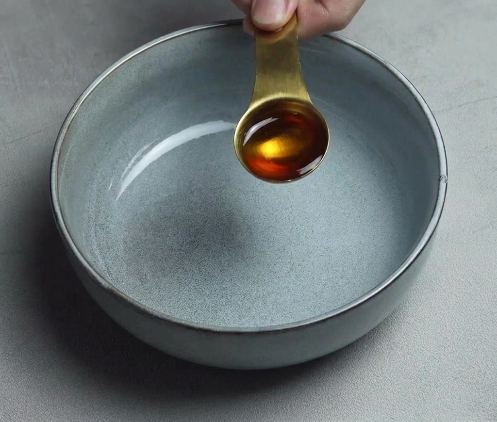 きれいな空のグレーのセラミック製のラーメン鉢の底に、大さじ1杯の黄金色のごま油を注いでいる様子。