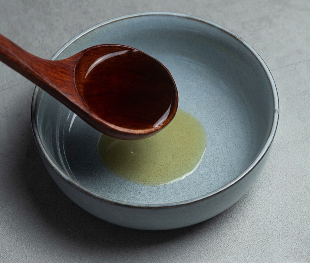 ごま油が入ったグレーのセラミック鉢に、木製のお玉で熱々の醤油ラーメンスープを注いでいる様子。
